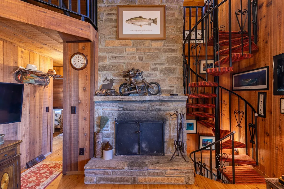 This view features the impressive stone fireplace, spiral staircase, and rustic decor with fishing memorabilia, evoking a classic mountain lodge feel. The scene suggests a perfect setting for warming up after outdoor activities or gathering on chilly evenings.