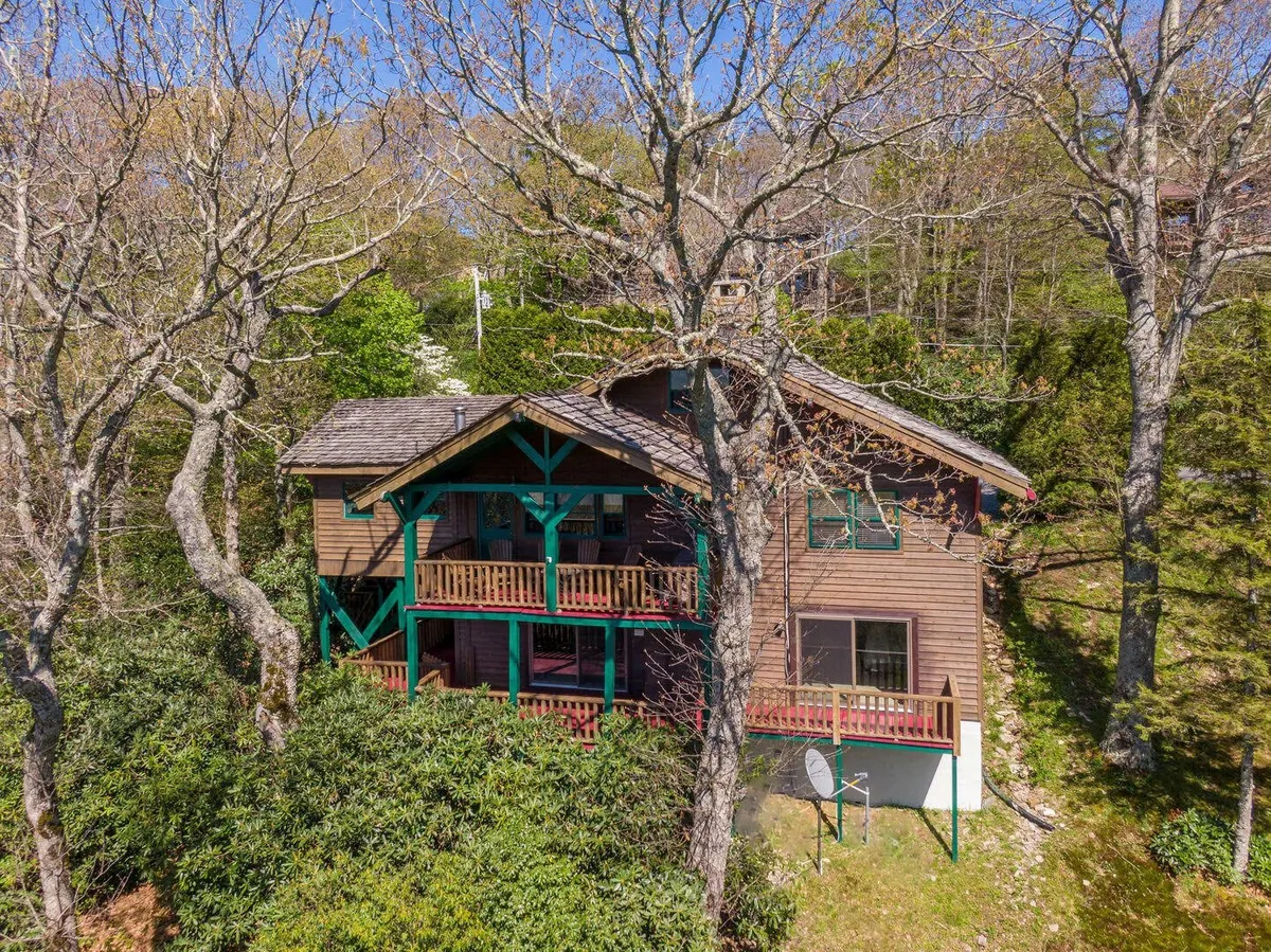 A tranquil exterior view of the home's wooden deck, surrounded by trees and featuring comfortable seating with a direct line to sunset views over the mountains. The green accents on the house and the peaceful setting invite relaxation outdoors.