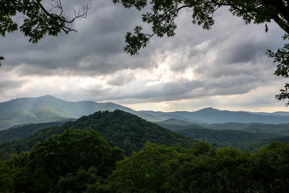 The image showcases a breathtaking, expansive view of the Blue Ridge Mountains from the property, with lush greenery in the foreground and dramatic clouds accentuating the distant peaks. The tranquil scene hints at sunrise or sunset, offering an inviting, peaceful atmosphere that highlights the natural beauty surrounding the home.