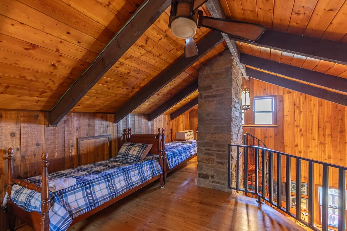 A warmly lit room with inviting plaid twin beds set under rustic wooden beams, a stone pillar, and a window providing night sky views. The space feels intimate and perfect for memorable sleepovers or relaxing mornings.