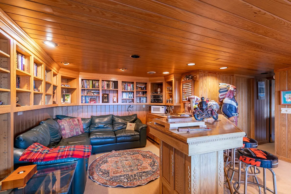 A comfortable corner of the entertainment space with a leather sectional sofa, wooden bar counter, bookshelves filled with books, and decorative accents. The arrangement encourages both intimate conversations and group relaxation, complementing the active game zone.