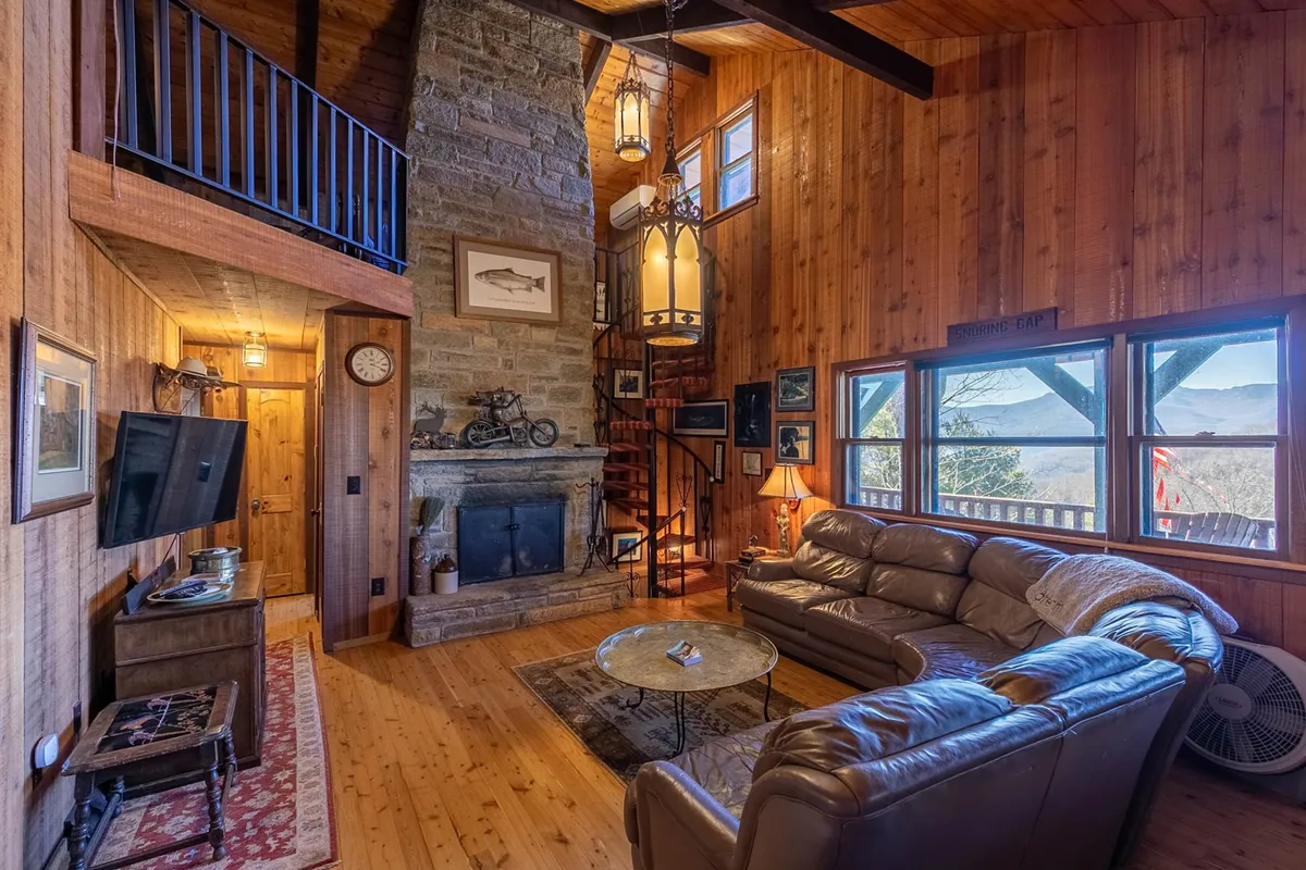 A warm, inviting living room featuring a dramatic two-story stone fireplace, plush leather sofa, spiral staircase, and expansive windows that flood the space with natural light and offer gorgeous mountain views. The scene captures a relaxed moment by the fire, with the mountains visible just beyond the glass.