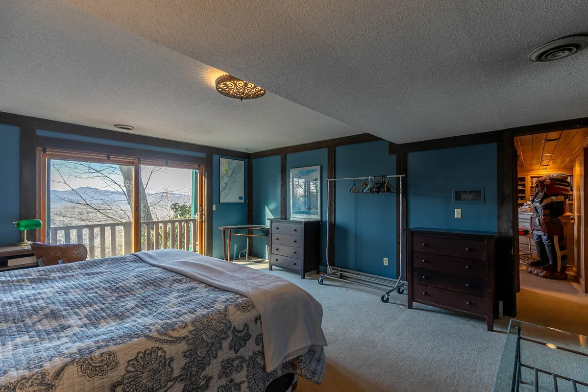 This image features a bed with patterned bedding positioned beside large windows that overlook an expansive mountain view. The wooden furniture and serene decor create a cozy yet connected-to-nature atmosphere, and the abundant natural light streaming in subtly emphasizes the mountain setting.