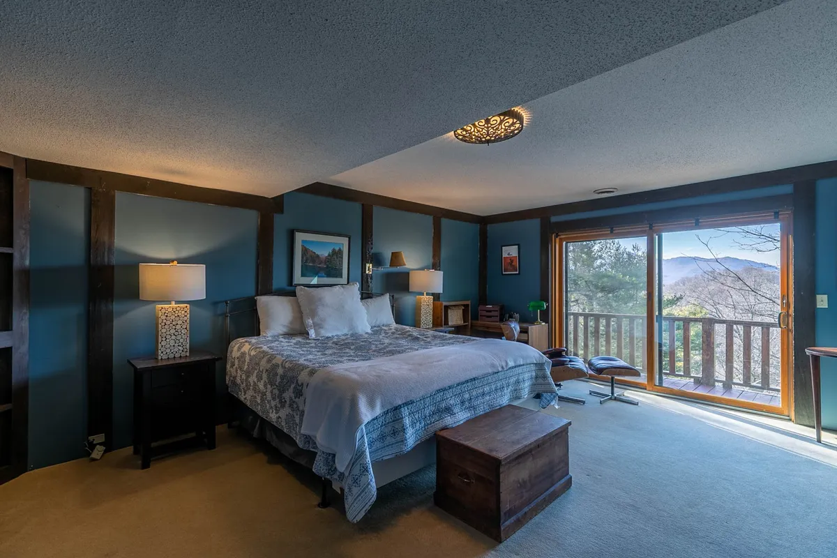 A serene bedroom with a comfortable bed positioned near a large window, allowing natural light to flood the space. A nightstand with a lamp and tasteful artwork provide a cozy, inviting atmosphere. The mountain view is visible through the window, reflecting the promise of waking up to Grandfather Mountain vistas.