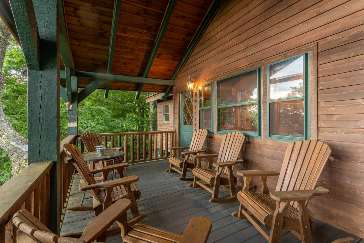 A welcoming porch scene featuring classic wooden rocking chairs, a sturdy railing, a small table with mugs, and an exterior light fixture. The porch overlooks surrounding nature, offering a tranquil space to relax and socialize outdoors.