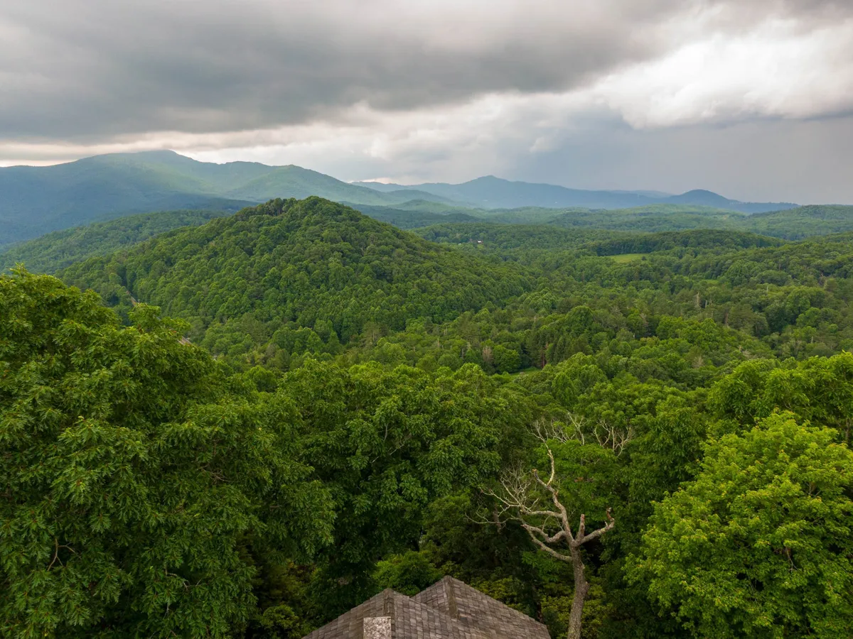 A sweeping view of rolling hills, dense forests, and a majestic mountain range under a cloudy sky. This wide landscape is vibrant and full of natural beauty, ideal for hikes, photography, and taking in breathtaking vistas.