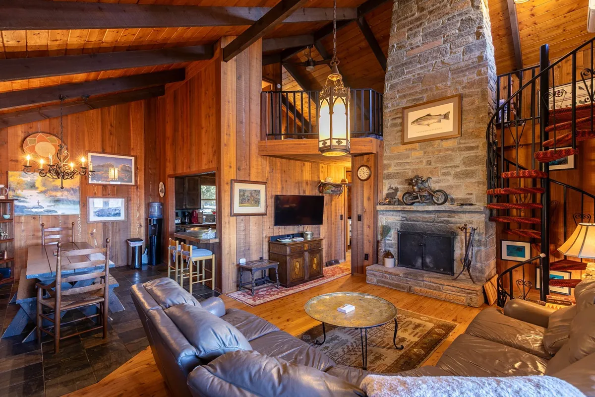 This image showcases a spacious living area centered around a grand stone fireplace, with a spiral staircase and a dining table visible. The room is bathed in warm light, evoking a welcoming, cozy atmosphere that highlights communal gatherings and relaxation after outdoor activities.