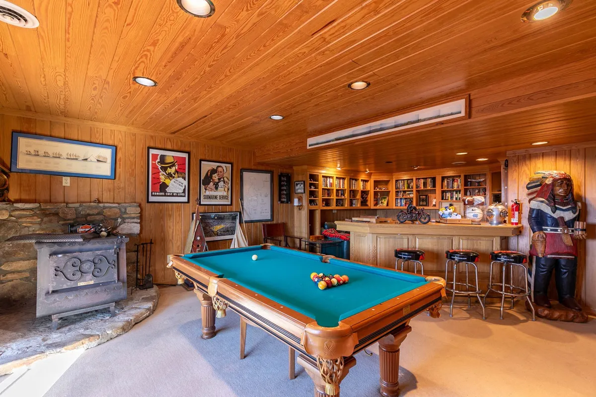 This image captures a lively game room setting, with a pool table at the forefront, a built-in bar area in the background, a cozy fireplace, decorative statues, and framed artwork adorning the walls. The lighting and furniture create an inviting and sociable atmosphere, ideal for group gatherings or friendly competitions.