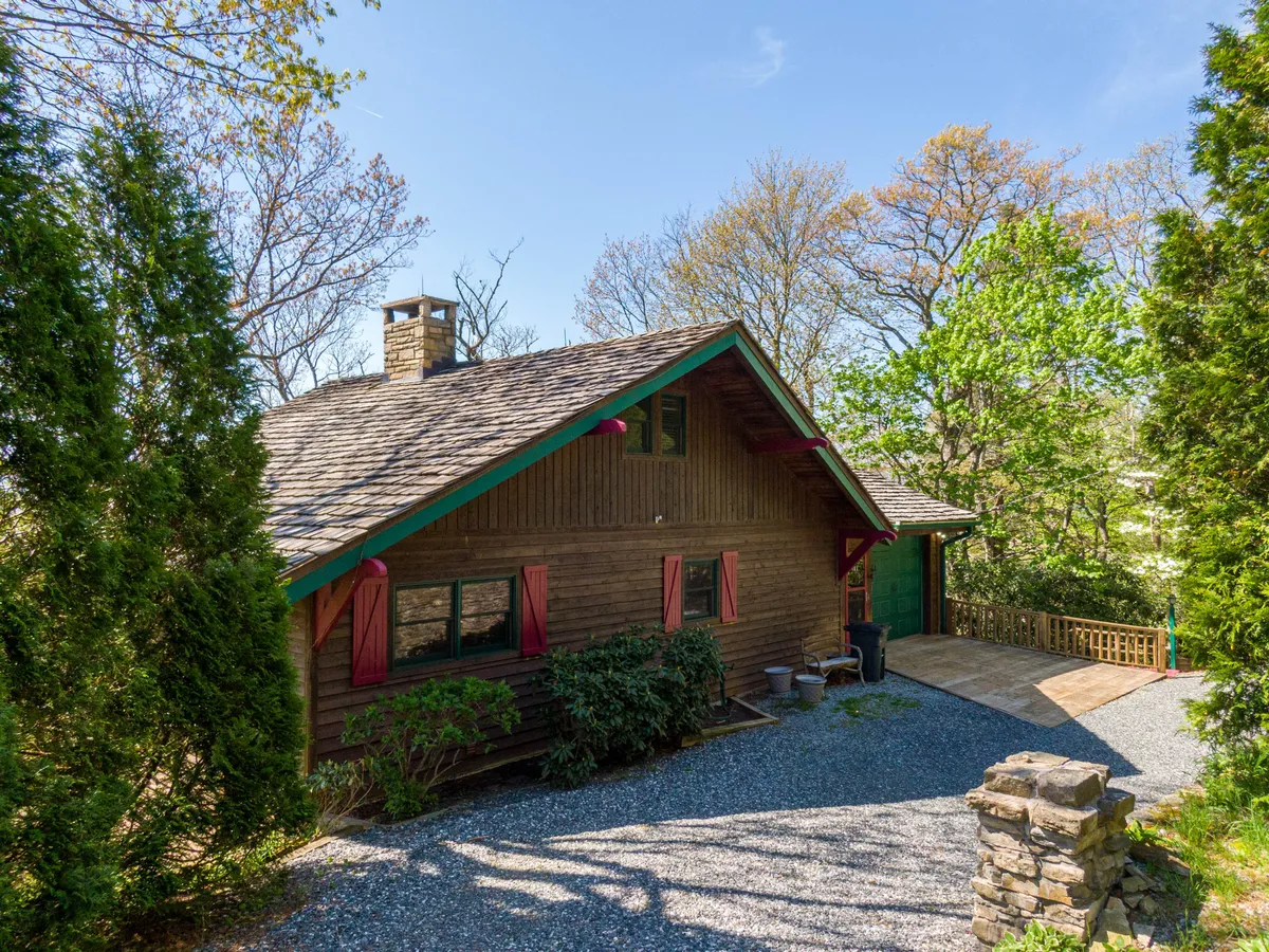 A cozy wooden cabin with a green roof and a prominent stone chimney sits nestled among tall trees. The porch offers inviting seating and the surrounding natural setting promises tranquility and immersion in nature. The image captures the cabin's exterior and its seamless integration with the landscape.