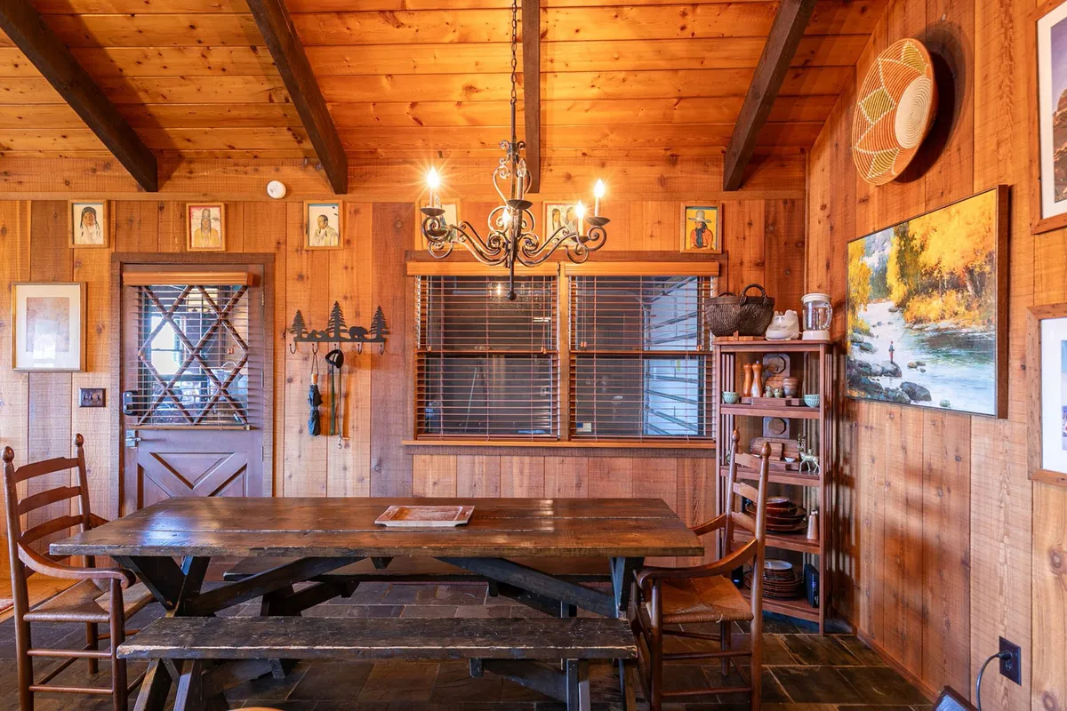 This image features a large wooden dining table, surrounded by chairs beneath a decorative chandelier. The backdrop includes artwork on the walls and slatted window blinds, suggesting a communal space ideal for gatherings, meals, and game nights. The warm, cozy lighting invites togetherness and conversation.
